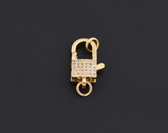 May include: A gold-colored clasp with a rectangular, gem-studded center. The clasp has a hinged top and a small ring at the bottom. The background is a solid black.