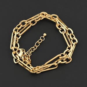 May include: A gold-coloured chain bracelet with a layered design. The bracelet features elongated rectangular links and a smaller chain for adjustable sizing. The clasp is visible, and the overall style is elegant and modern.