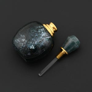 Op de afbeelding: Een decoratieve parfumfles met een donkere, gemarmerde uitstraling. De fles heeft een goudkleurige dop en een bijpassende stopper met een groene edelsteen top. De stopper is apart, waardoor de heldere glazen applicator zichtbaar is.