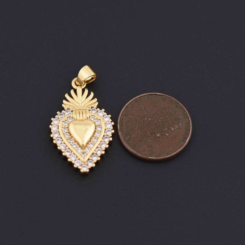 May include: A gold-colored pendant in the shape of a heart, adorned with clear crystals around the edges. The pendant has a small loop at the top for a chain. A penny is next to the pendant for size comparison.