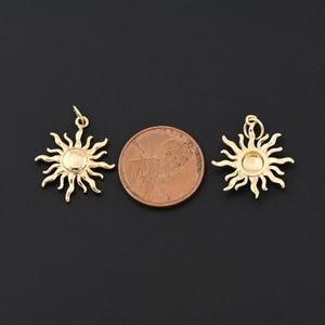 May include: Two gold-colored sun charms with a penny for size comparison. Each charm features a central disc surrounded by stylized rays. The charms have a loop at the top for hanging. The background is a solid black.