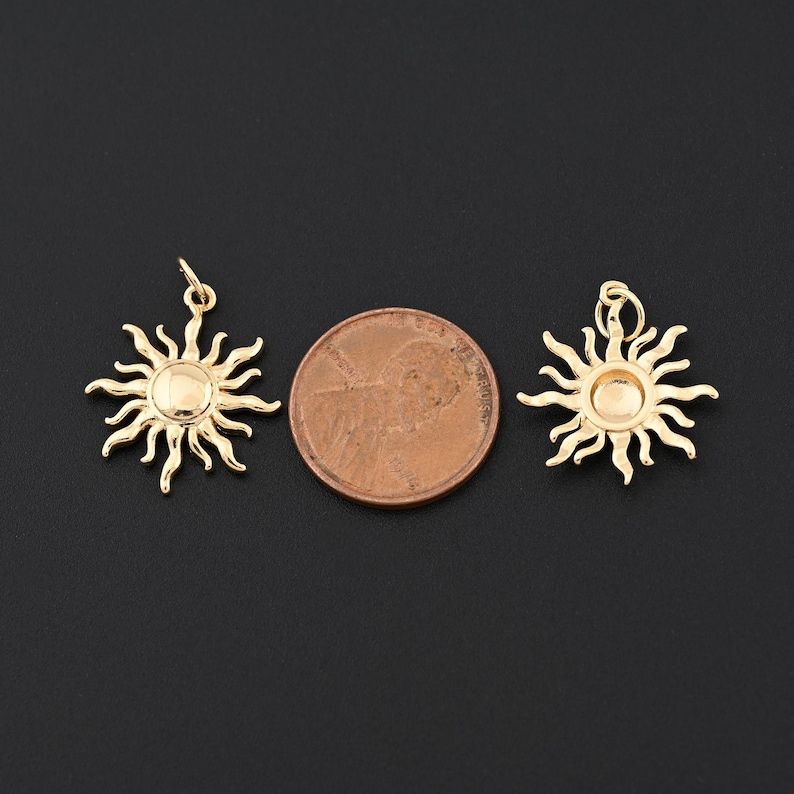 May include: Two gold-colored sun charms with a penny for size comparison. Each charm features a central disc surrounded by stylized rays. The charms have a loop at the top for hanging. The background is a solid black.