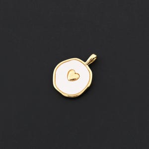 Puede incluir: Un colgante dorado con un centro de esmalte blanco y un coraz&oacute;n dorado. El colgante tiene una forma redondeada e irregular con un peque&ntilde;o lazo en la parte superior para una cadena. El colgante es probablemente para joyer&iacute;a.