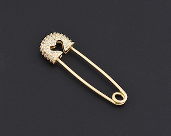 May include: A gold-colored safety pin with a heart-shaped cutout in the pin's head. The head of the pin is encrusted with small, clear crystals. The pin is set against a black background.