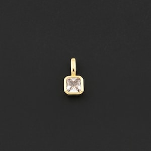 May include: A gold-colored pendant with a square, light pink gemstone. The gemstone is set in a gold bezel and has a small loop at the top for a chain. The pendant is displayed against a black background.