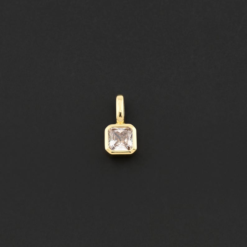 May include: A gold-colored pendant with a square, light pink gemstone. The gemstone is set in a gold bezel and has a small loop at the top for a chain. The pendant is displayed against a black background.