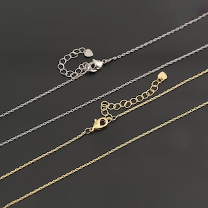 Pode incluir: Duas correntes metálicas delicadas, uma em tom prateado e outra em tom dourado, com um design de elos finos. Ambas incluem um fecho lagosta, uma secção de extensão ajustável com elos circulares e um pequeno pendente em forma de coração.