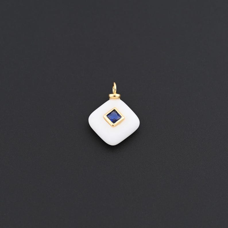 May include: A white, diamond-shaped pendant with a gold bail. The pendant features a square, deep blue gemstone set in the centre. The pendant is set against a dark background.