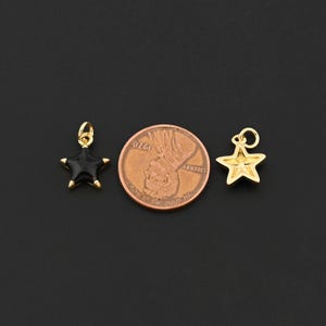 May include: Two star-shaped charms. One is black enamel with gold-colored trim, and the other is gold-colored. A penny is between the charms. The charms have a loop for hanging. The background is black.