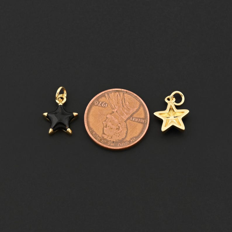 May include: Two star-shaped charms. One is black enamel with gold-colored trim, and the other is gold-colored. A penny is between the charms. The charms have a loop for hanging. The background is black.