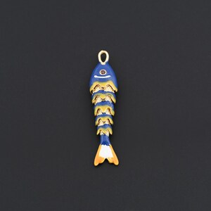 以下が含まれることがあります： 金と黄色のアクセントが付いた青い魚の形をしたペンダント。ペンダントの上部近くには小さな赤い宝石があり、吊り下げ用の金のループが付いています。尾はオレンジと白です。魚の長さは約7.6cmです。