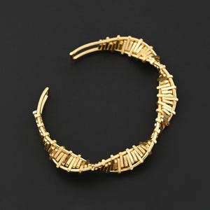 May include: A gold-colored cuff bracelet with a woven design. The bracelet features a series of rectangular bars woven together, creating a textured surface. The open design allows for easy wear. The bracelet is set against a dark background.