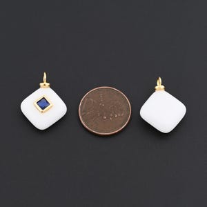 May include: Two white, diamond-shaped pendants with gold-coloured bails. One pendant features a square, blue gemstone. A penny is next to the pendants for size comparison. The pendants are on a black background.