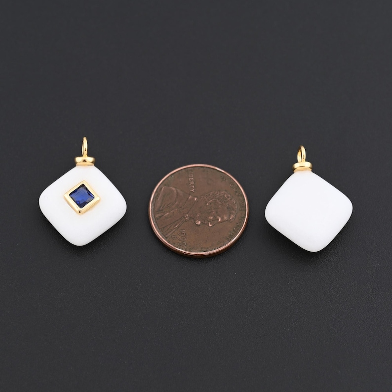 May include: Two white, diamond-shaped pendants with gold-coloured bails. One pendant features a square, blue gemstone. A penny is next to the pendants for size comparison. The pendants are on a black background.