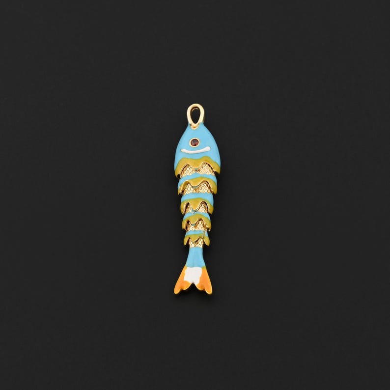 May include: A fish-shaped pendant featuring a gold-tone metal skeleton layered with light blue enamel sections. The head includes a small dark red circular accent, and the tail is finished with a white and orange enamel tip.