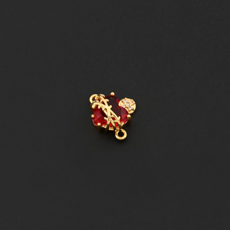 May include: A gold-colored charm with a red gemstone heart shape, accented with a smaller, round, clear-stone cluster. The charm has a loop at the top and bottom for attachment. The design is set against a black background.