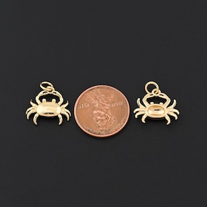 May include: Two gold-colored crab charms with a penny for size comparison. The charms have a loop at the top for hanging and are detailed with crab features. The background is a solid black.