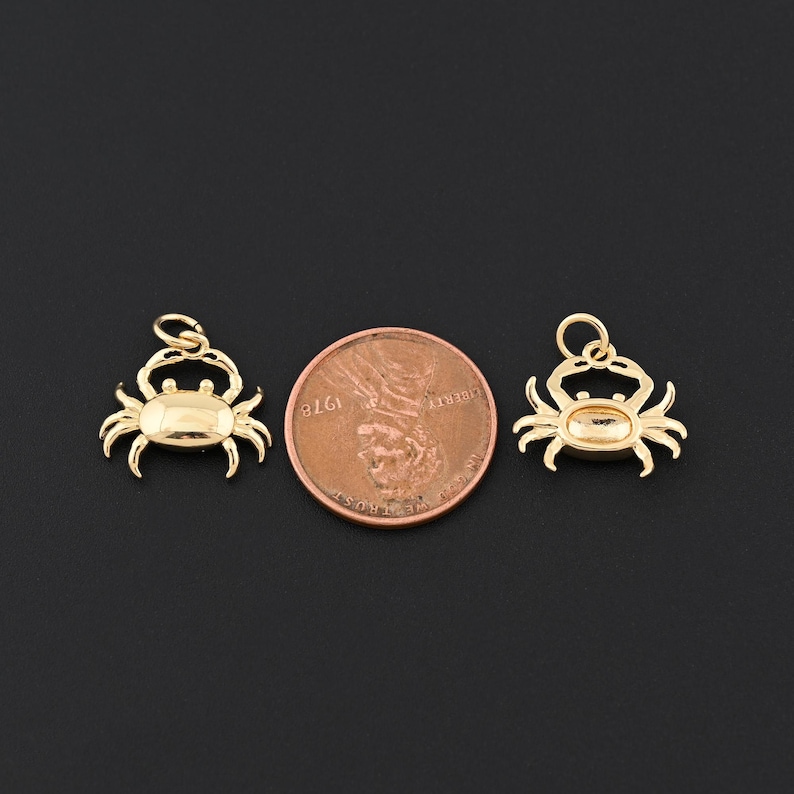 May include: Two gold-colored crab charms with a penny for size comparison. The charms have a loop at the top for hanging and are detailed with crab features. The background is a solid black.