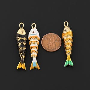 May include: Three metal fish-shaped pendants with textured scales and small loops at the top. One is black and gold, one is white, orange, and blue, and one is yellow and green. A copper-colored US penny is placed between the pendants.