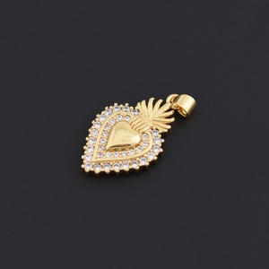 May include: Gold-colored heart pendant with a halo of small, clear crystals. The pendant features a heart shape in the center, surrounded by the crystals. A small loop is at the top for a chain. The pendant has a decorative design.