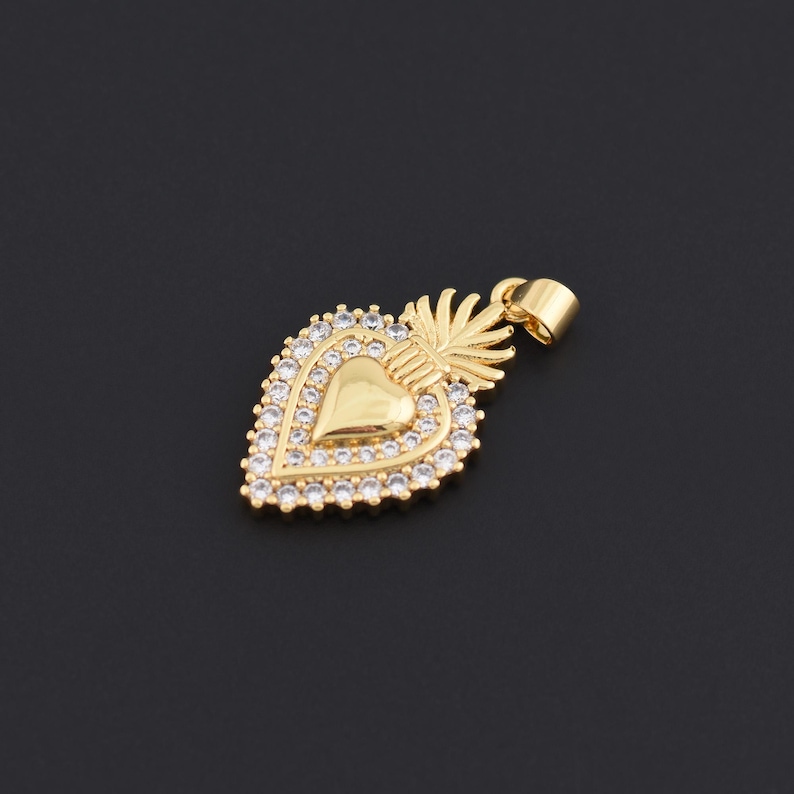 May include: Gold-colored heart pendant with a halo of small, clear crystals. The pendant features a heart shape in the center, surrounded by the crystals. A small loop is at the top for a chain. The pendant has a decorative design.