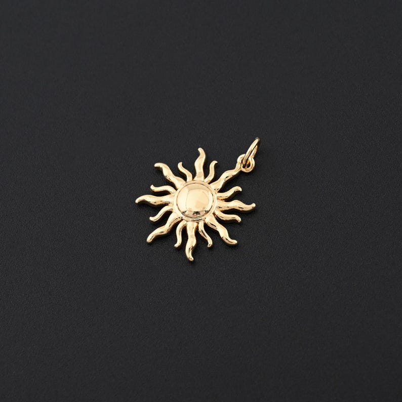 May include: A gold-colored sun pendant with a loop for a necklace. The sun has a circular center and multiple rays extending outwards. The pendant is set against a black background, highlighting its shape and metallic finish. The sun pendant is a jewelry item.