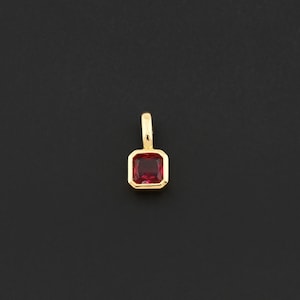 May include: A gold pendant with a rectangular, deep red gemstone. The gemstone is set in a gold bezel, and the pendant has a small loop at the top for a chain. The background is a solid black.