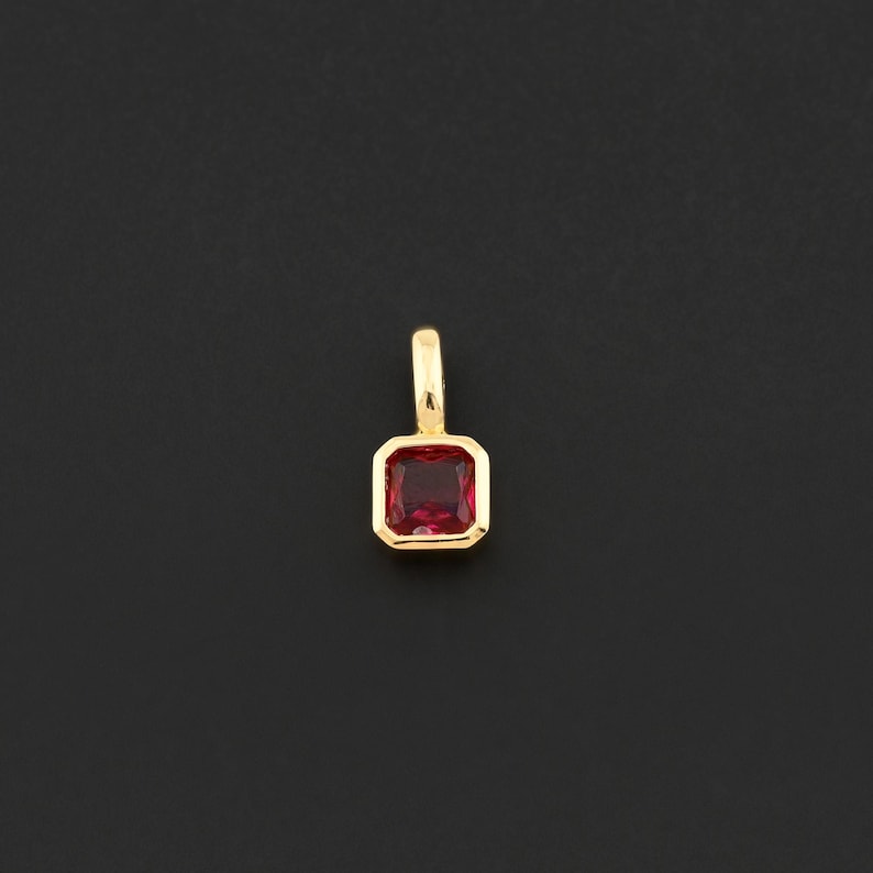 May include: A gold pendant with a rectangular, deep red gemstone. The gemstone is set in a gold bezel, and the pendant has a small loop at the top for a chain. The background is a solid black.