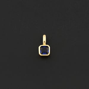 May include: A gold pendant with a square, deep blue gemstone. The gemstone is set within a gold frame, and the pendant has a small loop at the top for a chain. The background is a solid black.