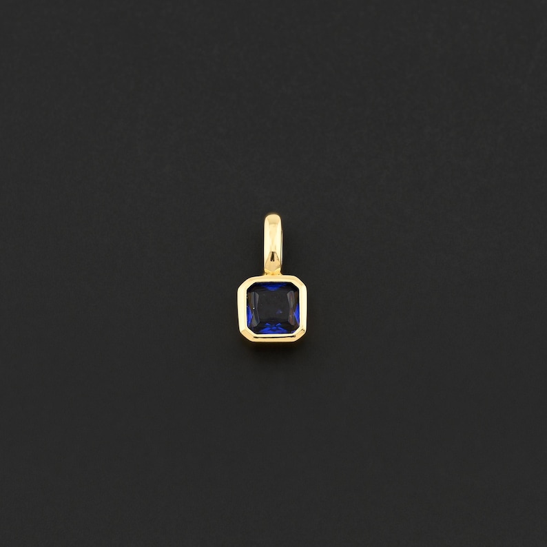 May include: A gold pendant with a square, deep blue gemstone. The gemstone is set within a gold frame, and the pendant has a small loop at the top for a chain. The background is a solid black.