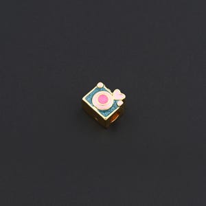 May include: A gold-toned charm in the shape of a camera. The camera has a square body with teal enamel, a pink lens, and a pink heart-shaped detail. The charm is likely for a bracelet or necklace.
