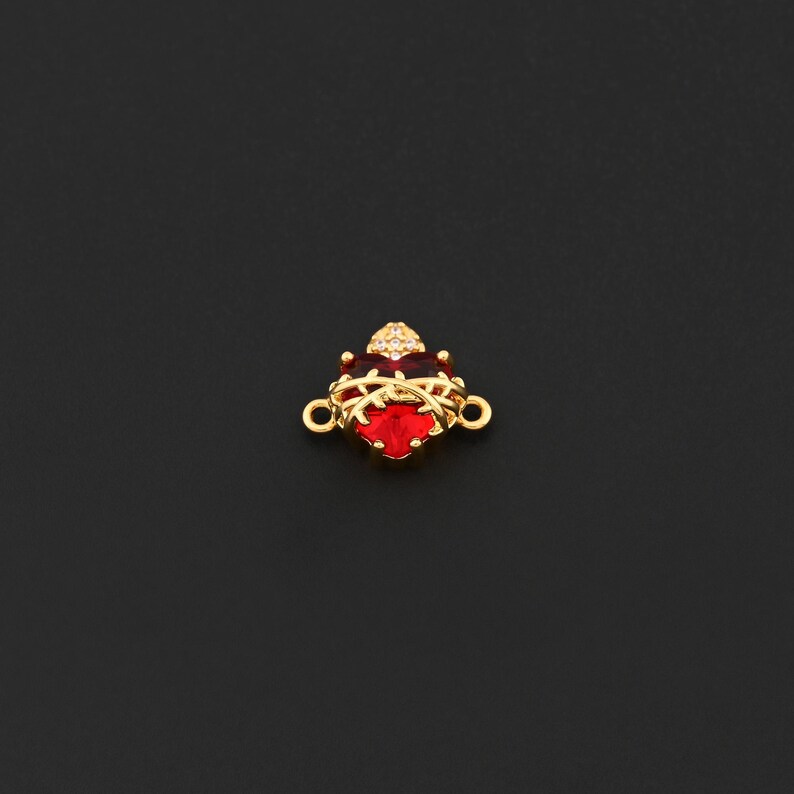 May include: A gold-colored charm with a red heart-shaped center stone. The charm has a crown-like design with small clear stones and two loops for attachment. The background is black.