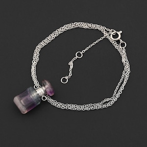 Puede incluir: Un collar de plata con un pequeño colgante de frasco de perfume. El frasco es de piedra translúcida morada y transparente con un tapón a juego. El collar tiene una cadena delicada y un cierre de broche, sobre un fondo oscuro.