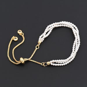 Puede incluir: Una pulsera dorada con tres hilos de perlas blancas. La pulsera presenta una delicada cadena y una cuenta deslizante para ajustar la talla. El diseño incluye pequeñas gemas transparentes en los extremos de la cadena, sobre un fondo oscuro.