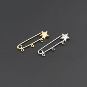 May include: Two safety pins, one gold-toned and one silver-toned, each featuring a five-pointed star at the head and two small circular loops attached to the pin shaft for hanging charms or other decorative elements on the metal structure.