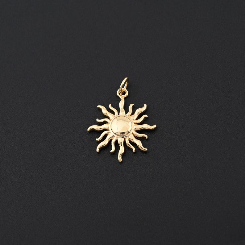 May include: A gold-colored sun pendant with a detailed design. The pendant features a central circular element surrounded by stylized rays. The pendant is suspended from a small loop, ready to be attached to a chain or necklace. The background is a solid black.
