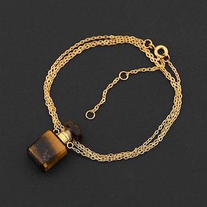 Puede incluir: Un collar dorado con un pequeño colgante rectangular. El colgante presenta tonos marrones y dorados, similares al ojo de tigre. El collar tiene un diseño de cadena delicada y un cierre de broche, sobre un fondo oscuro.