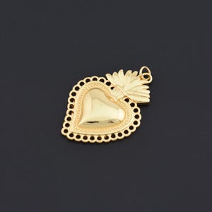 May include: A gold-colored heart-shaped pendant with a floral design and a small loop for a chain. The pendant has a detailed, textured border and a smooth, polished heart shape. The overall design is ornate and decorative.