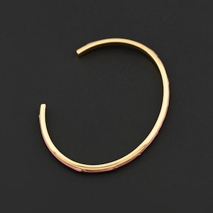 Könnte beinhalten: Ein goldfarbenes Armreif-Armband mit rosa Emaille-Inlay. Das offene Armband hat eine glatte, polierte Oberfläche und ein zartes Design. Die rosa Emaille ist auf der Innenseite des Armbands sichtbar.