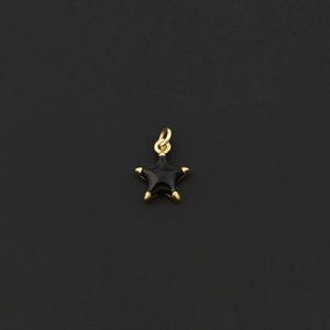 May include: A small, star-shaped pendant with a black enamel finish and gold-colored accents. The pendant has a loop at the top for attaching to a chain or bracelet. The star's points are tipped with gold, and the background is a solid black.