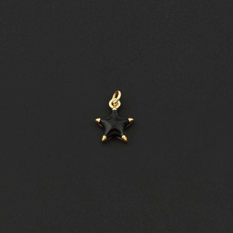 May include: A small, star-shaped pendant with a black enamel finish and gold-colored accents. The pendant has a loop at the top for attaching to a chain or bracelet. The star's points are tipped with gold, and the background is a solid black.