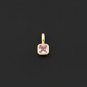 May include: A gold-colored pendant with a square, light pink gemstone. The gemstone is set in a gold bezel and has a small loop at the top for a chain. The pendant is displayed against a black background.