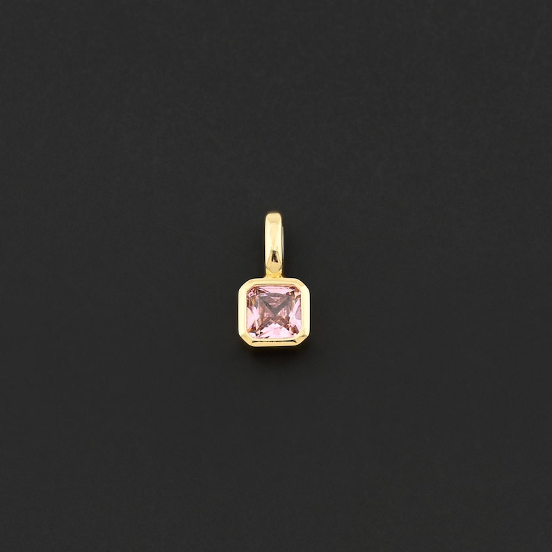 May include: A gold-colored pendant with a square, light pink gemstone. The gemstone is set in a gold bezel and has a small loop at the top for a chain. The pendant is displayed against a black background.