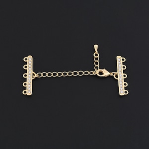 Puede incluir: Cierre de joyería dorado con una cadena y dos barras. Cada barra presenta una fila de pequeños cristales transparentes. El cierre tiene un cierre de mosquetón y múltiples bucles para la fijación. El fondo es negro.