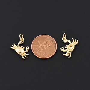 Puede incluir: Dos colgantes dorados con forma de cangrejo, con una moneda para comparar el tama&ntilde;o. Los colgantes tienen un dise&ntilde;o detallado, incluyendo pinzas, patas y un cuerpo texturizado. Sobre un fondo negro.