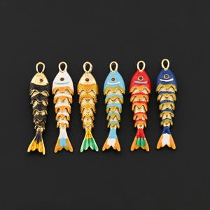 May include: Six colorful fish-shaped charms with gold-tone metal accents. Each charm features a different color scheme, including black, white, gold, blue, red, and a mix of colors. The charms have a loop at the top for attaching to jewelry or other crafts.