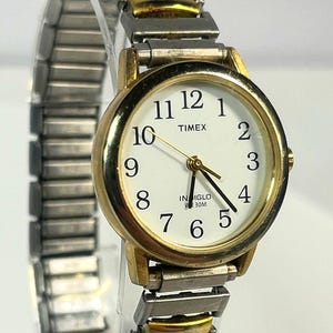 May include: A Timex wristwatch with a gold-tone bezel and a silver and gold-tone band. The watch face is white with black numerals and hands. The watch has the word "TIMEX" printed on the face.