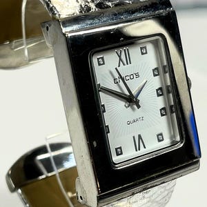 May include: Silver-toned watch with a rectangular face and a textured band. The watch face is white with black hands, Roman numeral hour markers, and the word "QUARTZ". The brand name "CHICO'S" is printed on the watch face.
