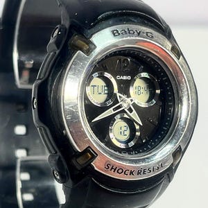 May include: Black Baby-G Casio watch with a silver bezel and black dial. The watch displays the day, time, and date. The watch band is black and made of a flexible material. The watch face has the words "Baby-G", "Casio", and "Shock Resist".