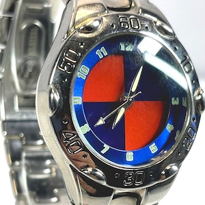 May include: A silver-toned wristwatch with a blue and red dial. The watch face features a blue outer ring with white numerals and a red and blue quadrant design. The watch has a silver band and a classic, sporty style.